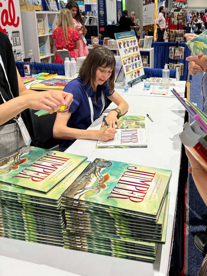 Photograph of Jessica Lanan sitting at a table in a coference events hall next to a stack of books, smiling and signing them for people who are standing in a line.