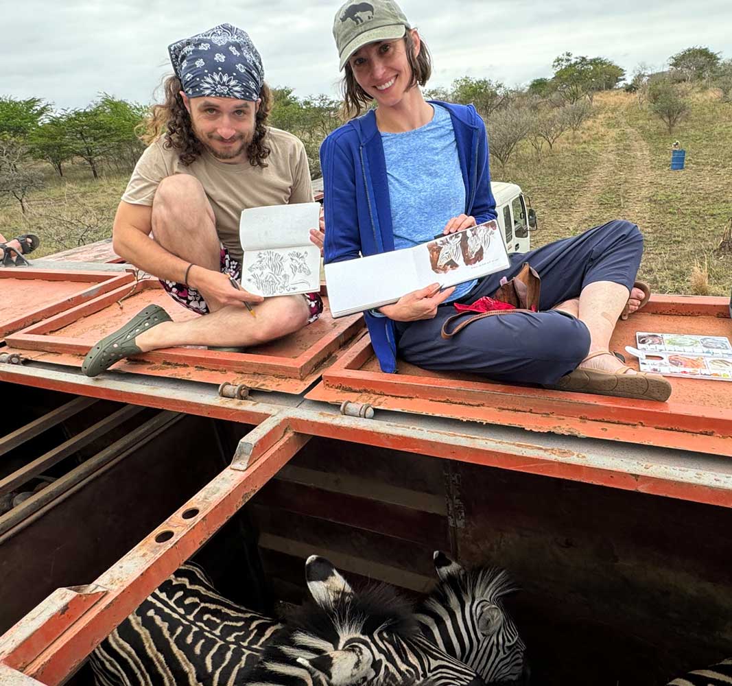 Photograph of author and illustrators Jessica lanan and Corbin Wilkin sitting on top of a container truck containing a herd of zebras. Both illustrators are holding up sketchbooks and smiling.