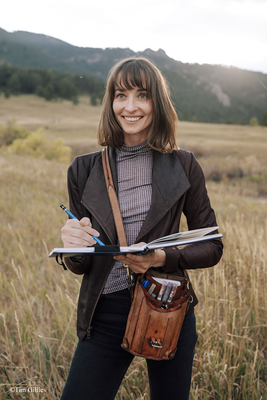 Photograph of Jessica Lanan sitting at her drawing table in her somewhat cluttered studio mixing paint on a watercolor palette, with paintbrushes and art supplies in the foreground